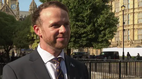 North Cornwall MP Ben Maguire stood outside Westminster in the sunshine. He is looking away from the camera with a smile. He is wearing a black suit and a white shirt with a Cornish flag pin badge.