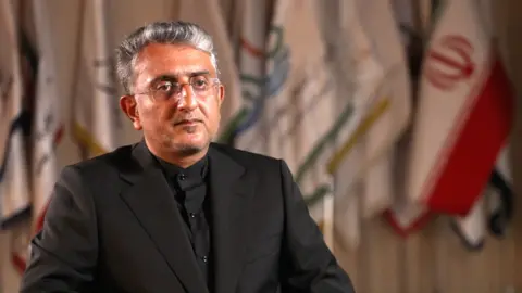 Jack Garland / BBC A man wearing a black shirt and jacket looks at the camera. He is sitting down with flags behind him, one of which is the Iranian flag. 