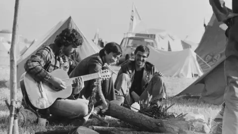 Chris Moorhouse/Evening Standard/Hulton Archive/Getty Images Three men sit down at a free festival in Glastonbury in 1974. It is a black and white picture and one of them is playing an acoustic guitar.