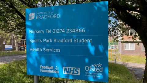 A blue sign with white lettering in the shade of a tree.
