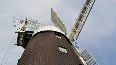 Wicken windmill in Wicken near Sohan in Cambridgeshire. The windmill has a brown exterior, reaching high into the sky. The picture has been taken from the ground and the white sails of the mill are stationery. The picture has been taken on a day with a blue sky and some overcast cloud. 