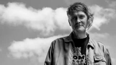 Alfie Harco A black and white image of a man with short fringe and longer sections swept to the side . He is standing outside with a cloudy sky visible behind him. He is wearing a jacket and a black t-shirt with white logo.