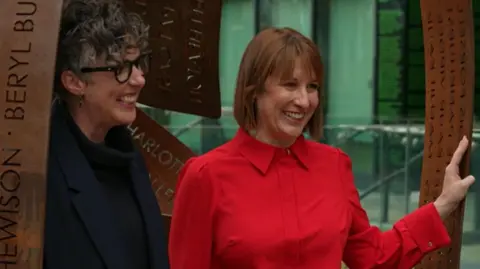BBC Rachel Reeves wears a red blouse and is smiling at the camera as she leans with one hand against the sculpture. Artist and creator Pippa Hales stands next to her. She has short curly grey hair and is smiling at the camera.