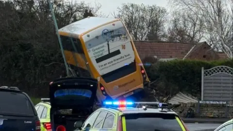 A yellow double decker Stagecoach bus that has crashed and come off the road. It is angled upwards and leaning against a pole. There is a crushed fence and brick wall near it. Two police cars can be seen near the bus.