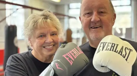 Simon Thake/BBC A couple who are in a gym together wearing black t-shirts and boxing gloves. They are smiling.