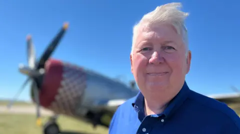 Richard Moseley is standing on a field which has a World War Two plane on it. He has short white hair and is wearing a blue shirt.