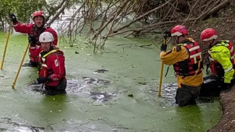 Essex County Fire and Rescue Service Firefighters in the water trying to get the sheep out of the water.
They are wearing helmets and their uniform and holding long sticks so they can help walk through the thick water 