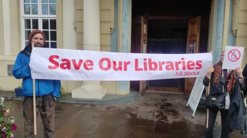 Campaigners outside the library protesting against the decision to re-locate the service. Two campaigners are holding a long white banner which says "Save our Libraries" in in red writing.