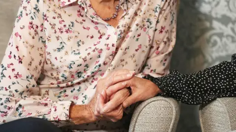 The image shows a close-up of two people sitting together, focusing on their hands. One person is wearing a light-coloured blouse with a floral pattern and a necklace, while the other is wearing a dark top with small white polka dots. The person in the floral blouse is gently holding the other person’s hand, which rests on the arm of a light upholstered chair. The background includes a patterned wallpaper in muted tones.