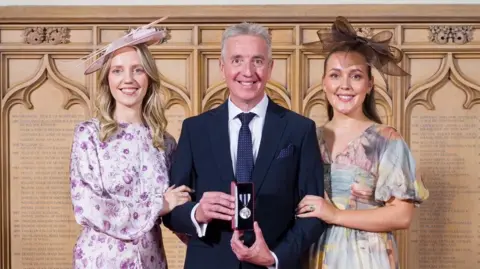 TVP Jim has short grey hair and is dressed smartly, whilst holding a medal. Each of his daughters cling on to his arms.