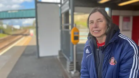 BBC Paula Mitchinson is smiling as she waits to board a train on Platform 1 at the Stadium of Light metro startion. She is wearing a navy blue Adidas jacket with Sunderland AFC's red and white crest. Her hair is short and grey. There is a green footbridge above the tracks.
