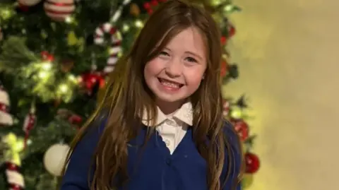 Nyla standing indoors in front of a decorated Christmas tree. The tree is adorned with warm white lights and a variety of festive ornaments, including red-and-white striped decorations, candy‑themed pieces, and round baubles. A sign reading “REINDEER WAY” is positioned prominently among the branches. Nyla is wearing a school uniform consisting of a blue cardigan, a white collared shirt, and a dark skirt, and is standing on a carpeted floor with hands clasped in front.