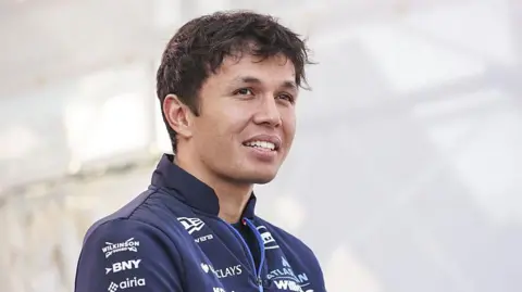 Getty Images Alex Albon looking into the distance past the camera in a closely cropped photo. He has short wavy dark hair and is wearing a navy blue Williams team zipped jacket. The photo is taken at the Japanese Grand Prix but the background behind him is just white.