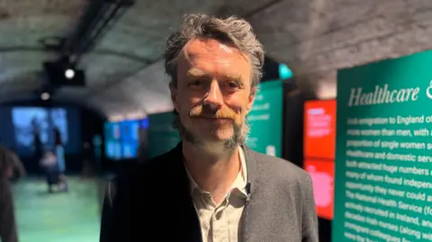 Man wearing a dark suit smiles at the camera. He is standing in front of an exhibition