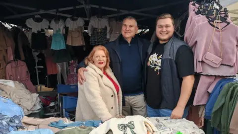 Emporium Productions Three people are on a stall with clothes above and below them. Smith in the centre has his arm around a long-serving stall holder, a woman, on the left and Smith's son, Joe, is on the right. They are looking at the camera.