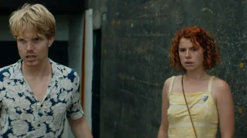 Escena al aire libre de la película Beast, filmada por Altitude Film Entertainment, donde se ve a dos personas de pie cerca de una pared de hormigón oscuro y la entrada de un edificio. El actor Charley Palmer Rothwell, con una camisa estampada de flores, se encuentra a la izquierda, mientras que la actriz Jessie Buckley, con una blusa sin mangas de color amarillo pálido, está a la derecha y lleva un bolso cruzado al hombro.