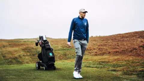Golfer Josh Simpson on the fairway of a golf course with his buggy behind him.