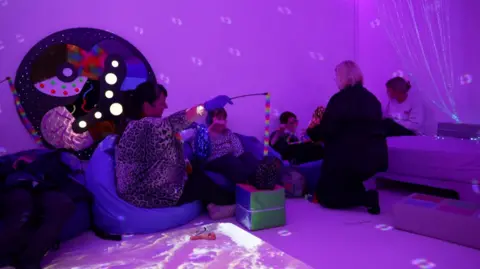 A group on people on bean bags in a sensory room with purple lighting and light projections of different shapes on the wall and floor