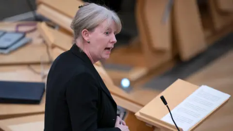 Shona Robison wearing a black jacket photographed from her right hand side while standing at a wooden lectern in the main debating chamber in the Scottish Parliament. She has blonde hair which is tied up.