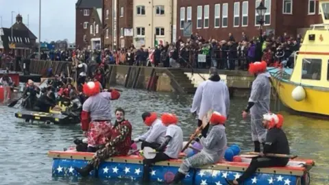 Poole Harbour Commissioners Crowds along a sea wall with people in fancy dress on the water aboard raft-type boats and bright yellow large boat to the right.