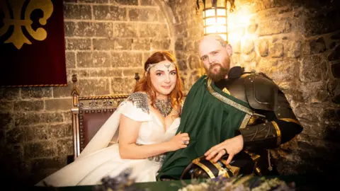 Seated portrait of two people in medieval-inspired attire inside a stone-walled room lit by a warm lantern. Jasmine who has ginger hair wears a white flowing dress with decorative silver shoulder pieces and a jewelled headpiece, while her husband wears dark armour with a green sash draped across the chest. They sit closely together on an ornate wooden chair beside a red banner with a gold emblem.