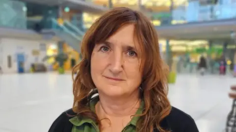 Paul Moseley/BBC Lucy Galvin is sitting in The Forum in Norwich looking at the camera. She has a black light cardigan over a green shirt.