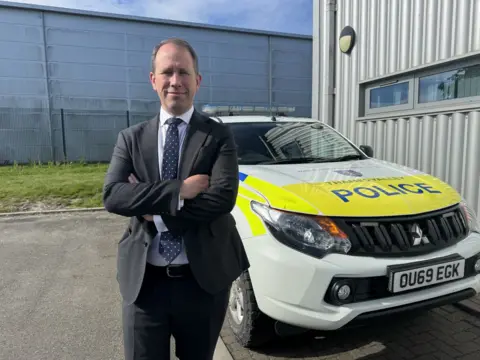 Dressed in a grey suit and tie Matthew Barber has his arms crossed in front of him. He's standing in front of a stationary police car which in turn is parked alongside a corrugated steel wall housing a police building in Wantage.