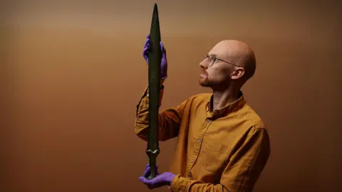Stewart Attwood Dr Matthew Knight is standing in a room holding the sword. Dr Knight has a bald head, a neatly trimmed beard and is wearing a mustard coloured shirt, and glasses. He is wearing purple, rubber gloves.