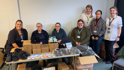 George King/BBC Seven women standing behind a table on which there are multiple boxes of slippers for elderly people.