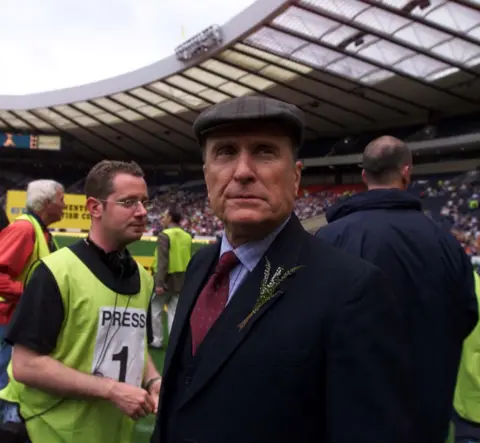 SNS Robert Duvall looks into the crowd while dressed as a Scottish football manager. There is a press photographer in the background.
