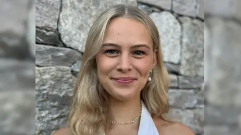 Lucia Shiner A woman with long blonde hair is smiling at the camera, she is wearing a gold necklace and a white halter neck top. She is stood in front of a stone wall. 