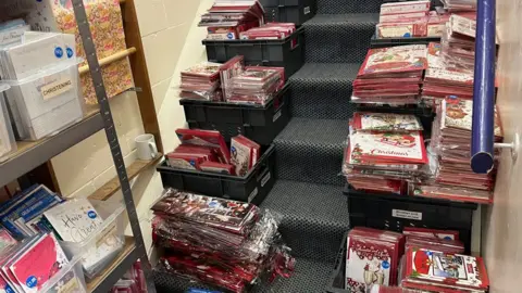 John Devine/BBC Piles of unsold Christmas card stock, stacked in black plastic boxes on the steps of a grey carpeted staircase, leaving a narrow walkway in the middle.