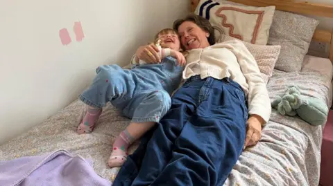 Hefina Everard Helen and Margot cuddle on a bed laughing. Margot has denim dungarees on, and Helen is wearing a white cardigan and denim trousers.
