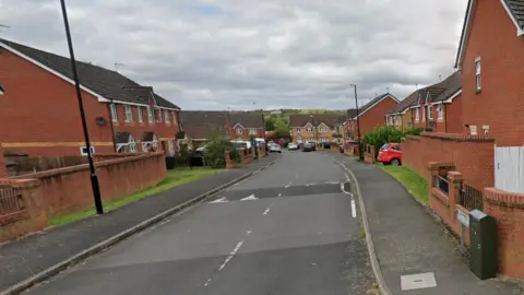 A wide image of Wolston Way in Coventry. Red brick homes line the street with cars parked in front of properties. There aer some grass verges visible with trees in the distance