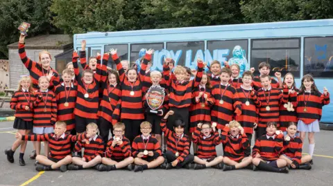 PA Media Rows of Year 6 pupils in matching red and black t-shirts modelled on Dennis the Menace. They are also wearing medals and have their hands in the air, cheering.