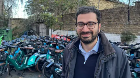 A man with a beard and glasses who is wearing a blue striped shirt beneath a blue top and grey-blue coat is standing in front of lots of e-bikes 
