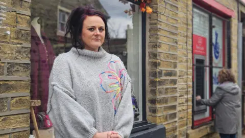 Aisha Iqbal/BBC A woman stands outside a shop front looking serious. She wears a grey jumper with a large floral pattern on it. In the background is a woman outside a post office, using a cash machine.