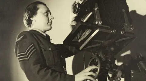 Sepia portrait of WAAF Sergeant Dorothy "Knicky" Chapman, standing in front of a large camera at RAF Iver Heath (Pinewood Studios). She is wearing her military uniform with sergeant's stripes on the sleeve.
