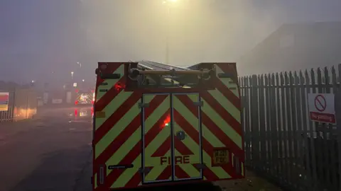 BBC The back of a fire engine parked next to a metal fence which has a sign saying 'no parking on it' with lots of smoke in front of it and the back of a second fire engine visible in the distance