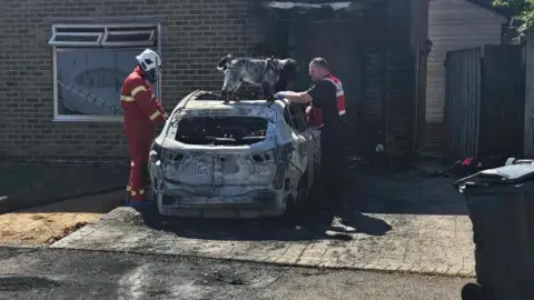 Emma Beci Two firefighters stand over a burnt out car