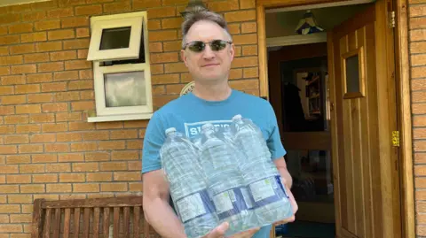 Kate Bradbrook/BBC A man in a blue T-shirt holding six large bottles of water wrapped in plastic packaging.