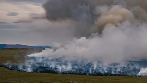 Aerial view of wide swathe of fire on charred moorland as smoke rises