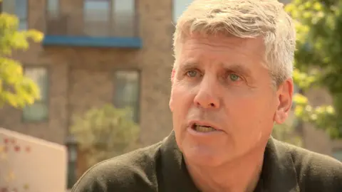 Man with short hair and black t-shirt looks directly at the camera with a serious expression.