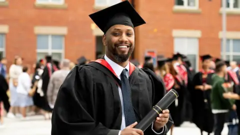 Hamza Hassan has a beard and is wearing full cap and gown and is holding a black scroll in front of other students in a university square.