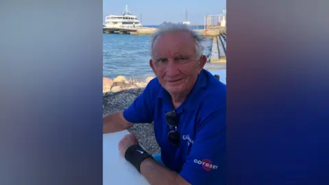 Merseyside Police A man in a blue polo shirt sit on a table in front of a harbour on a sunny day.