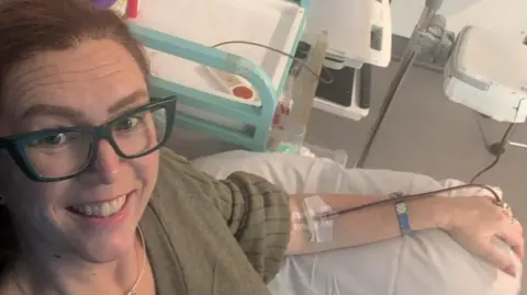 Sian Chathyoka Close up selfie of Sian smiling whilst laying in a hospital bed with a drip going into her left arm. She wears a green knitted jumper and has blue square glasses.