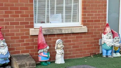 Kieran Simpson Image of several gnomes positioned on fake grass in front of a brick wall. In the background can be seen a window with a note stuck in it saying 'Gnomes available to take for free. P.S, please take them'. The gnomes all look very cheerful.