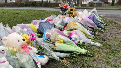 Jodie Halford/BBC Bunches of flowers are lying on grass at the side of a road. Some have messages written on them.