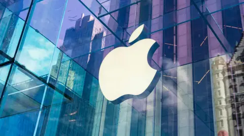 Getty Images The Apple logo on the side of a building. It is a cartoon of an apple with a bite taken out of it. The building is reflecting lots of colours from the surrounding area like purple, blue and green.
