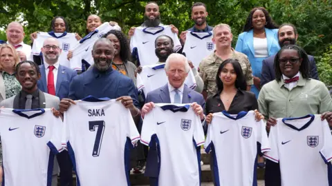 Reuters King Charles, Idris Elba, and a group of people stand holding up England football shirts